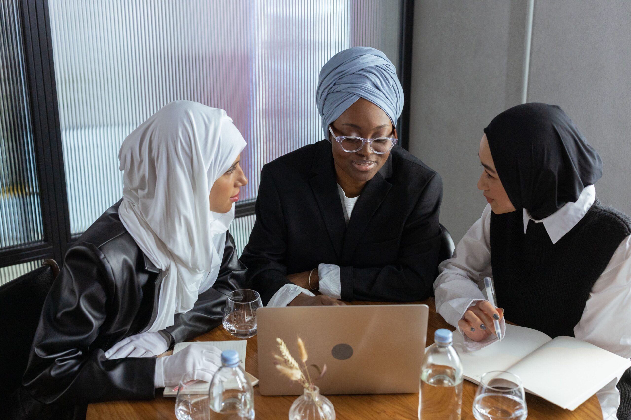 Three Muslim businesswomen in hijabs working together on a laptop in an office setting.